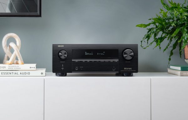Lifestyle image of Denon AVC-X3700H AV Receiver sitting on white AV cabinent next to books and a plant.