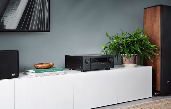 Lifestyle image of Denon AVC-X3700H AV Receiver on white AV cabinent with polk speakers connected, plant on the right and books on the left with bowl on top of the books.