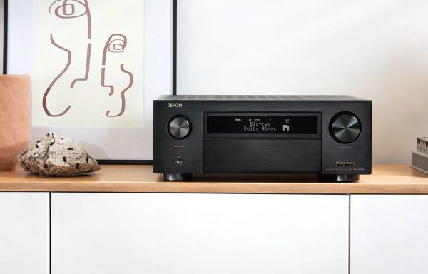 Lifestyle image of Denon AVC-X6700H AV Receiver sitting on cabinent powered on next to displays.