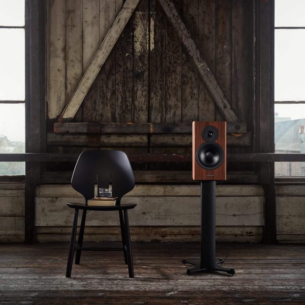 Dynaudio Emit 20 on speaker stand in lifestyle setting next to chair with book and glass on it.